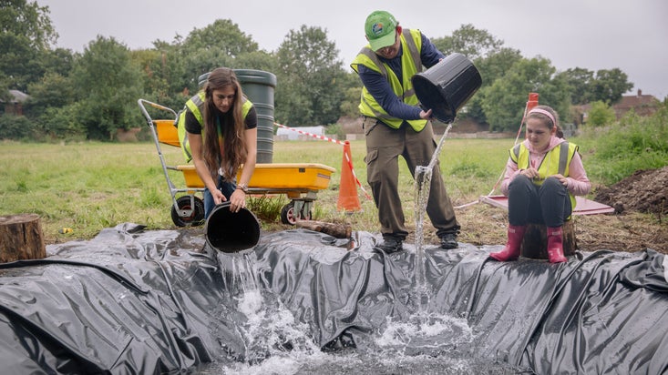 Three people pour buckets of water into a newly dug pond to create habitats for wildlife